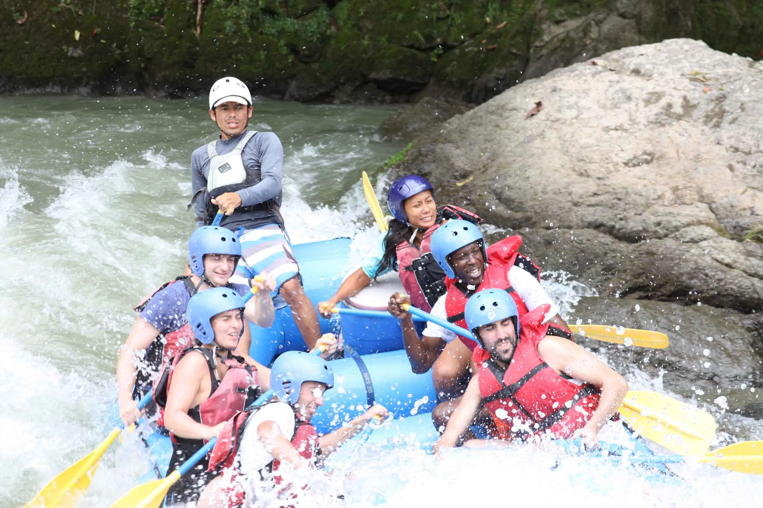 "Group of seven people experiencing high adrenaline while rafting, with excited expressions and splashing water around them."